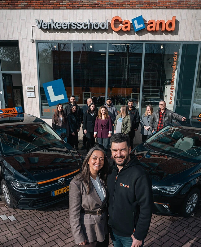 Team van Rijschool Caland in Amsterdam staat voor het pand van de rijschool, met twee lesauto’s op de voorgrond en het logo ‘Verkeersschool Caland’ zichtbaar op de gevel.
