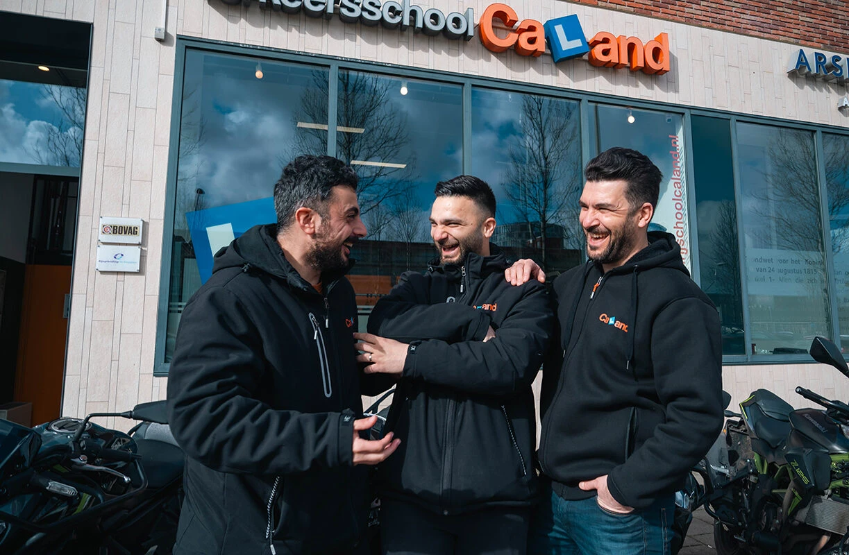 Drie instructeurs van Rijschool Caland staan lachend voor de vestiging in Amsterdam, een weerspiegeling van de kernwaarden kwaliteit, service en vertrouwen.